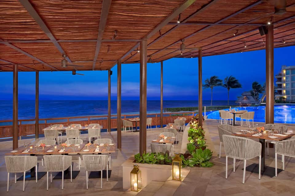 Beachfront dining pavilion at dusk with wooden pergola, white woven chairs, ocean view, and pool