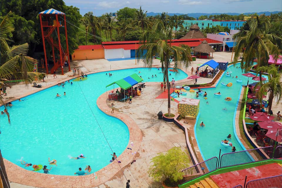 Large turquoise community pool with palm trees, water slide, and colorful resort buildings