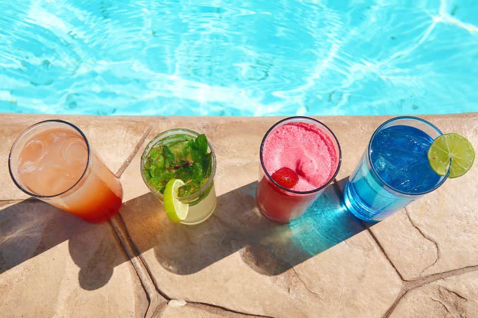 Four colorful cocktails on pool deck edge with turquoise water in background
