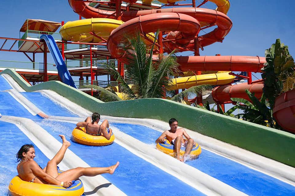 Water park guests sliding down colorful slides in yellow tubes with tropical palm trees and clear sky