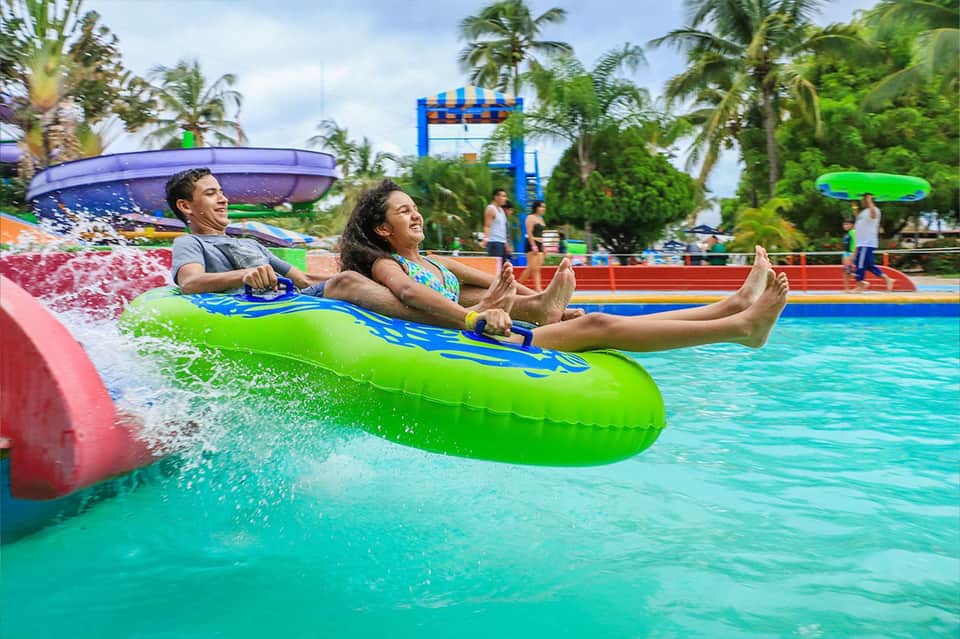Water park slide with family on green inflatable raft splashing into turquoise pool surrounded by palm trees