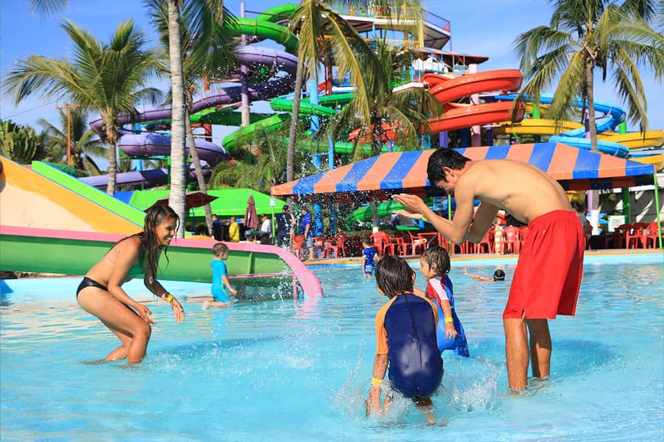 Children playing in shallow water park pool with colorful slides and palm trees