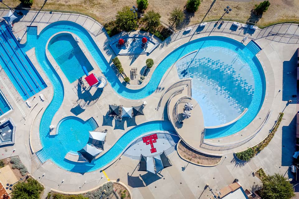 Aerial view of curved swimming pools and water park with umbrellas and landscaping