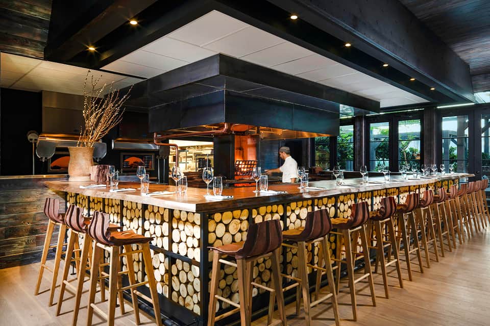 Contemporary hotel bar with long counter featuring geometric wood pattern, modern lighting, and open kitchen area