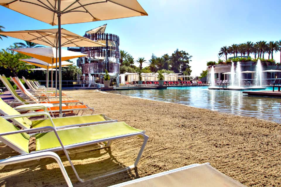 Colorful lounge chairs under umbrellas on a beach with fountain and palm trees in background