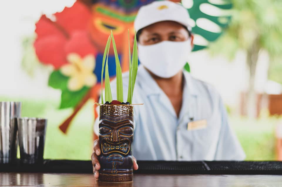 Bartender in white uniform holding tiki mug drink with colorful background and tropical flowers