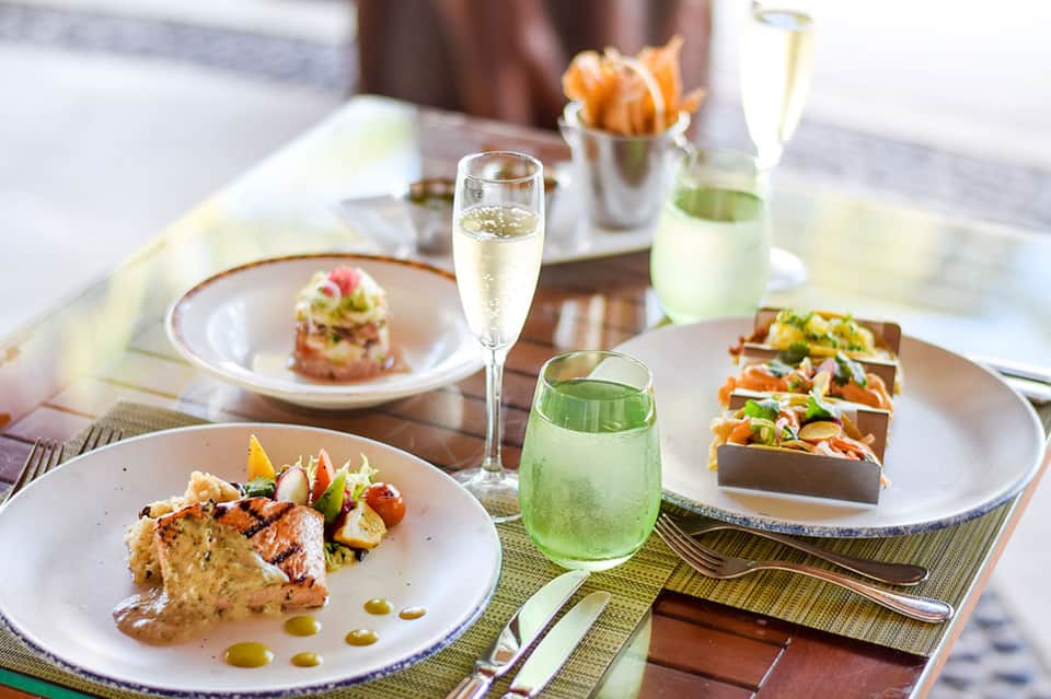 Fine dining plated seafood dishes with champagne, white wine, and green cocktail on beachside table