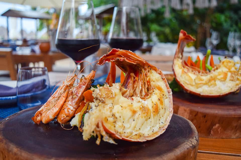 Grilled lobster tail with sauce on plate, wine glasses in background