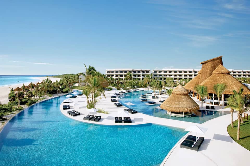 Large resort pool complex with thatched structures, white loungers, and beachfront location