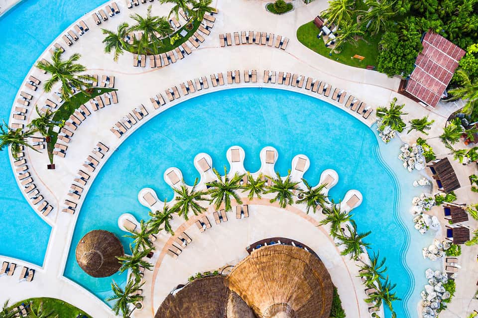 Overhead view of resort pools with loungers, palm trees, and shells in water