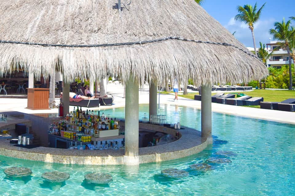 Overwater thatched-roof bar with turquoise lagoon water and resort buildings in background