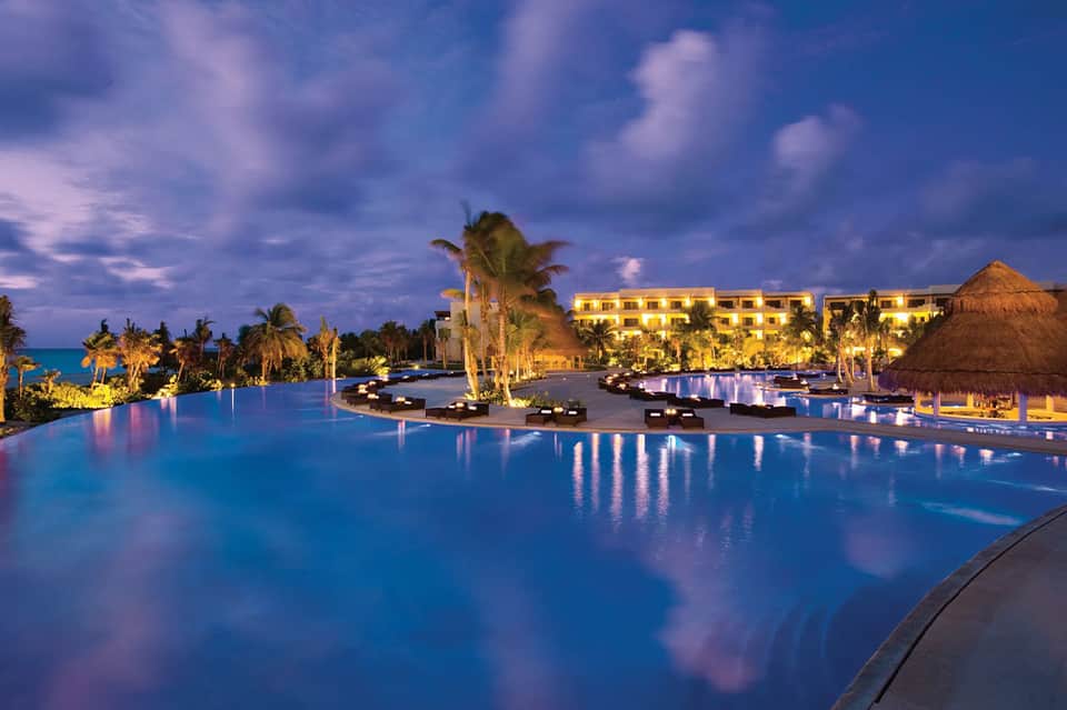Illuminated infinity pool at twilight with palm trees, lounge chairs, and resort buildings