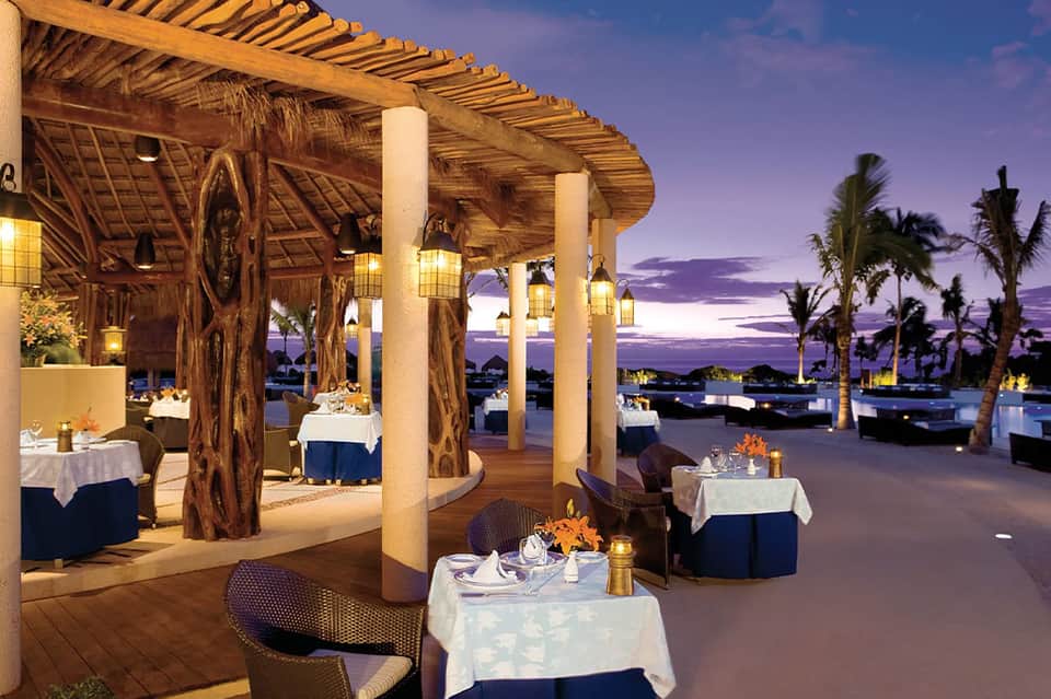 Upscale beachfront restaurant with teak pergola, white tablecloths, and purple twilight sky