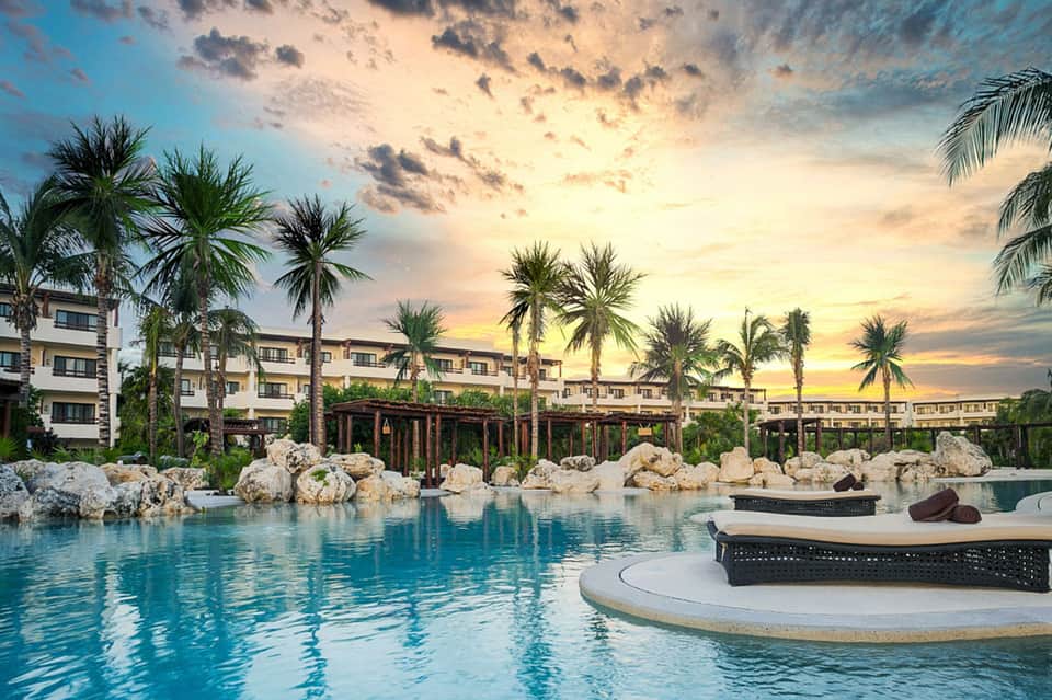 Resort pool at sunset with lounge chairs, palm trees, and buildings illuminated at dusk