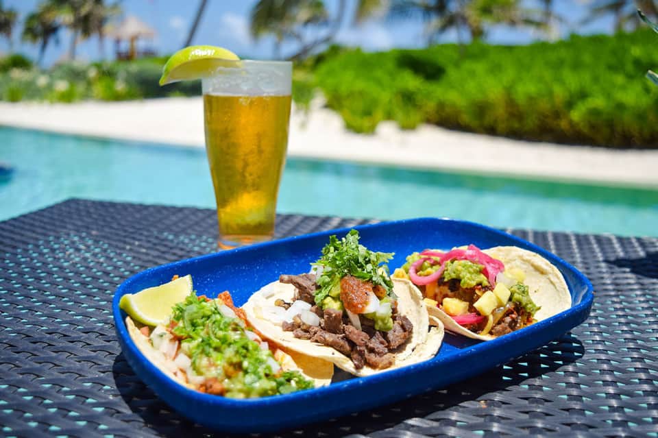 Three fresh tacos with lime on blue plate beside beer, poolside setting with ocean view