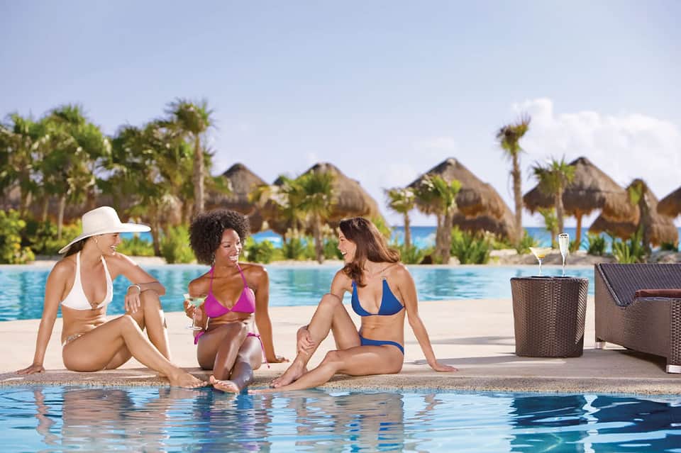 Three women in bikinis sitting poolside at a resort with thatched umbrellas in the background