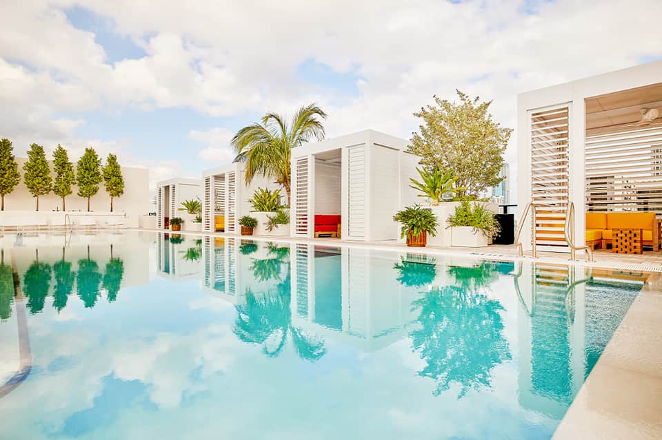 Modern resort infinity pool with white buildings, colorful lounge chairs, tropical landscaping, and reflective water surface