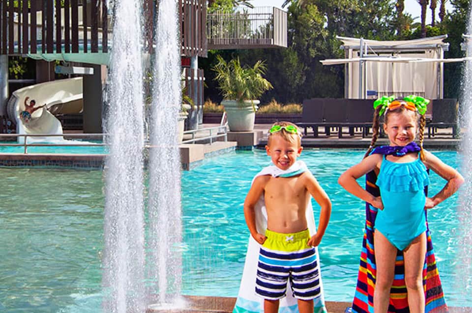 Two children in colorful swimwear posing by a resort pool with waterfalls and tropical landscaping.