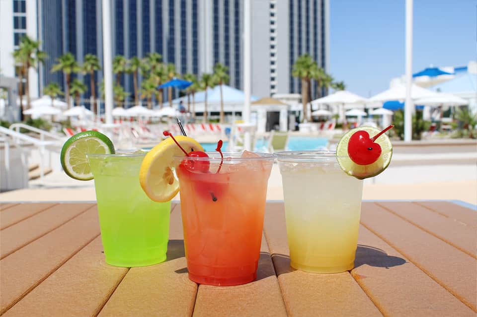 Three colorful tropical cocktails with fruit garnishes on deck overlooking resort pool and cityscape