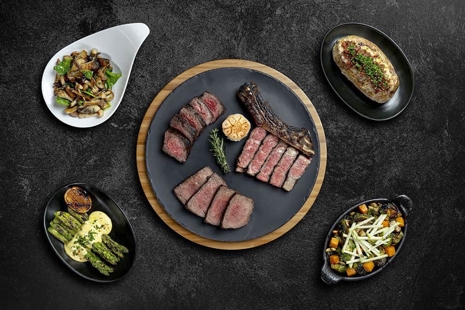 Gourmet plated steak dinner with sides of vegetables and garnishes on dark slate surface
