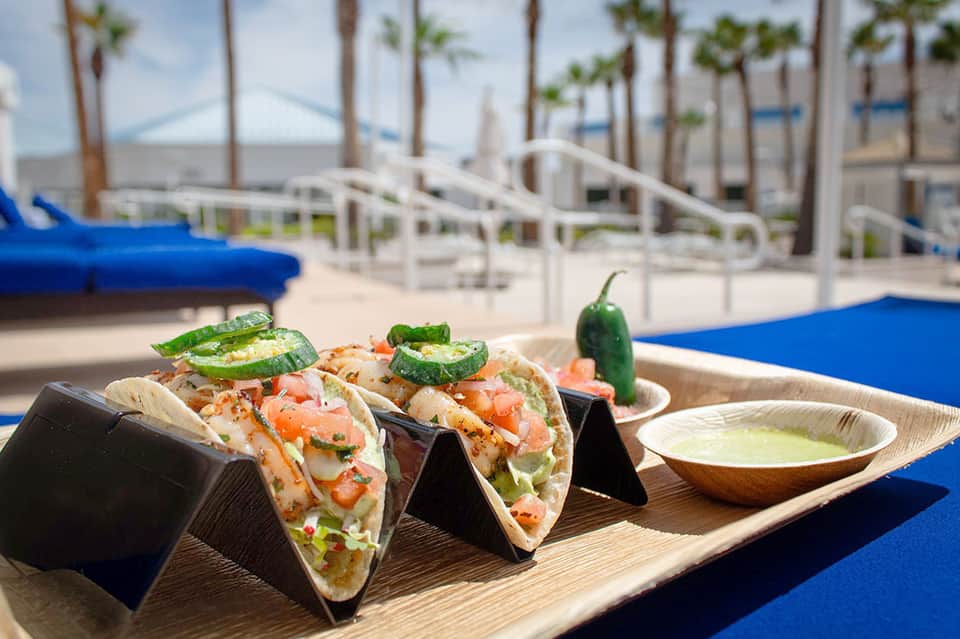 Three fish tacos with lime and jalapeño on wooden plate overlooking pool and ocean