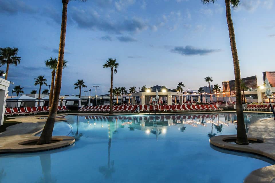 Large resort pool at dusk with palm trees, red lounge chairs, and illuminated buildings
