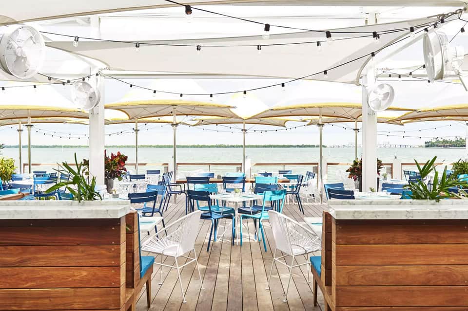 Waterfront restaurant under white retractable canopy with blue chairs, string lighting, and bay water views