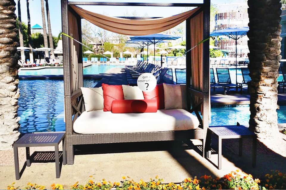 Luxurious poolside cabana bed with brown drapes, red and white pillows, overlooking resort pool and palm trees