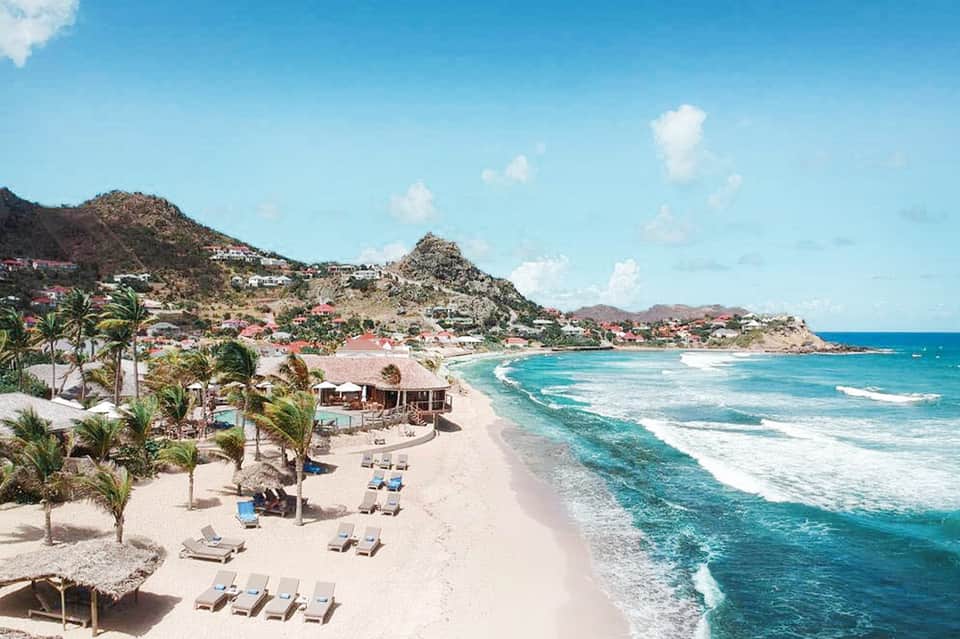 Scenic beach resort with mountainous backdrop, palm trees, lounge chairs, and turquoise ocean waves