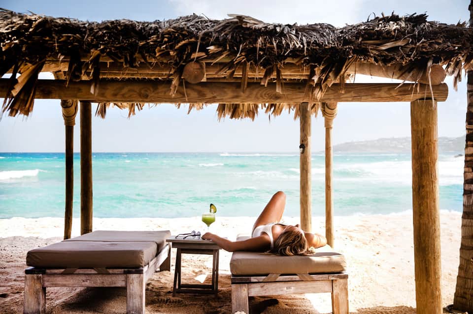 Woman relaxing on wooden lounge under thatched pergola overlooking turquoise ocean and beach