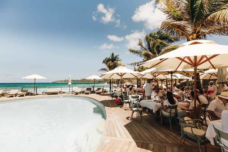 Beachfront restaurant with white umbrellas, palm trees, ocean view, and guests dining al fresco