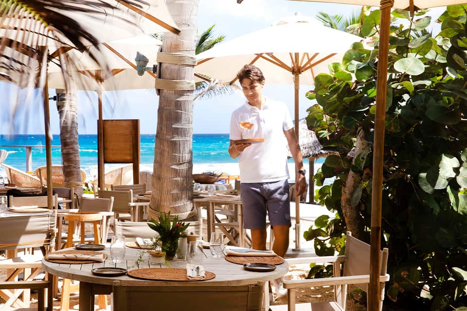Beachfront dining area with server, wooden posts, umbrellas, and turquoise ocean backdrop