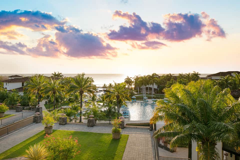 Aerial view of resort grounds with lagoon pool, palm trees, and ocean at sunset