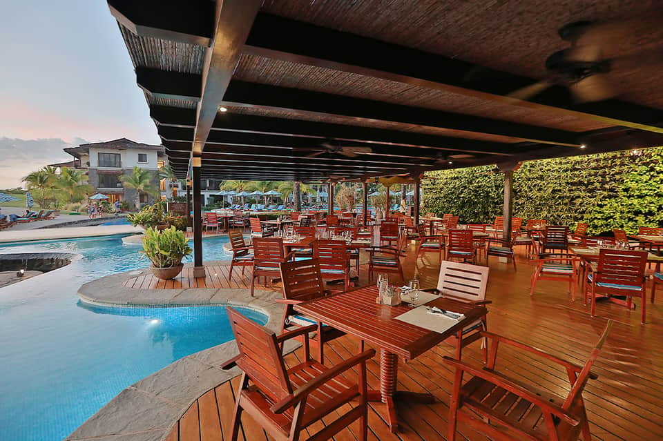 Covered poolside dining area with wooden furniture, tables, pool view, and lush landscaping