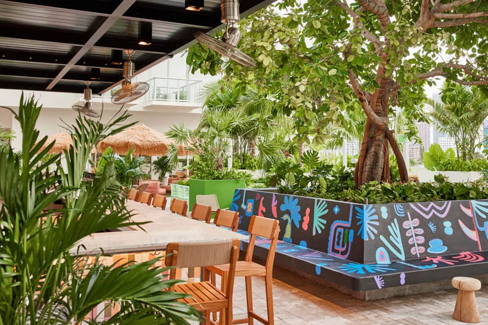 Tropical indoor dining area with colorful mural wall, palm plants, and thatched roof umbrellas