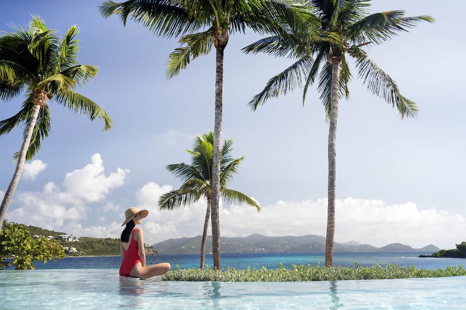 Woman in red dress relaxing in infinity pool with palm trees and tropical ocean view