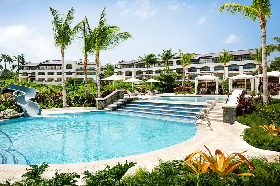 Resort pool complex with water slide, palm trees, lounge areas, and multi-story building