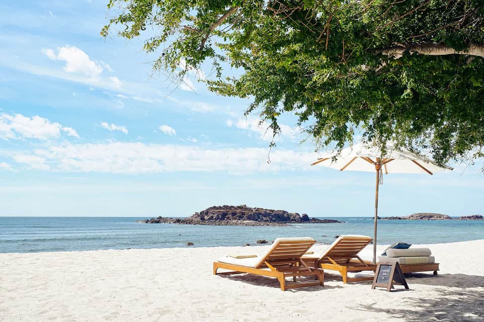 Beach lounge with wooden sunbeds, umbrella, tropical tree shade, and ocean view with small islands