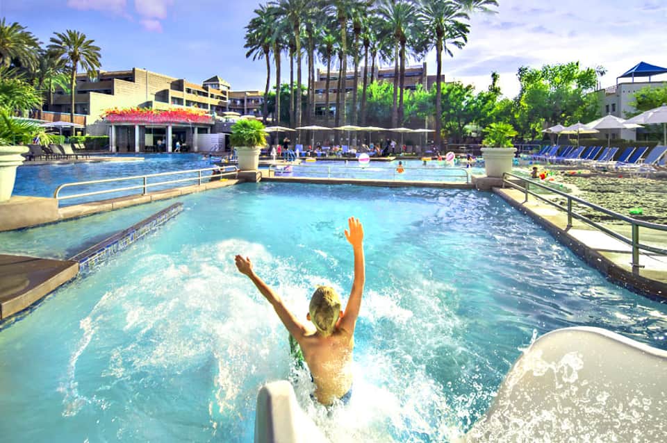 Young boy sliding into bright turquoise wave pool at resort with palm trees and multi-story hotel building