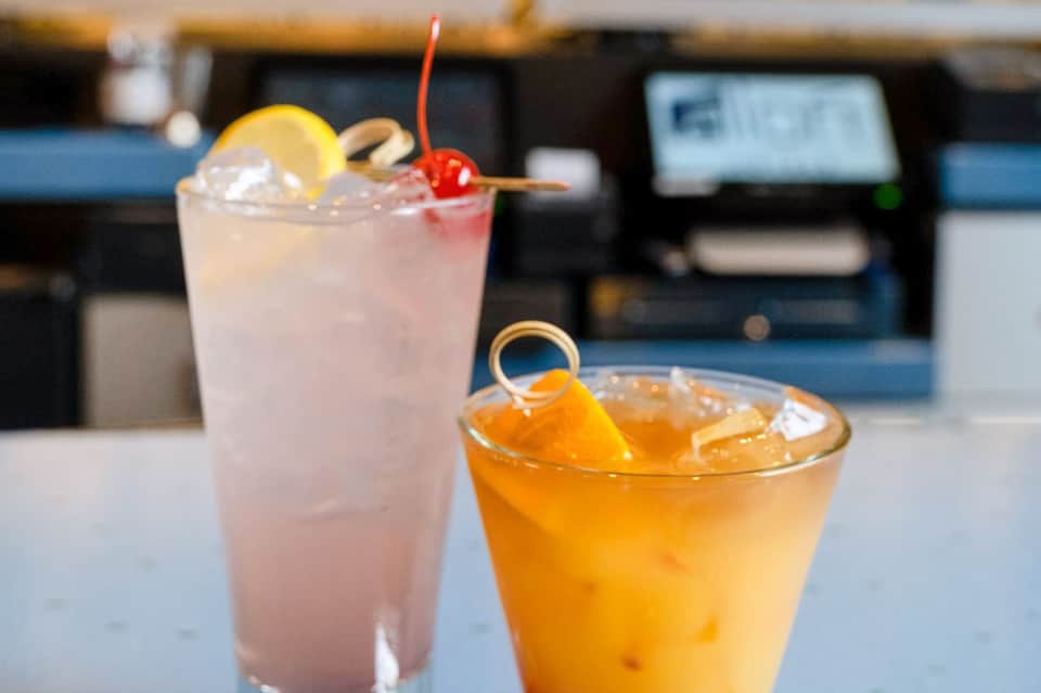 Two tropical cocktails at bar counter with lemon slice, cherry garnish, and blurred background