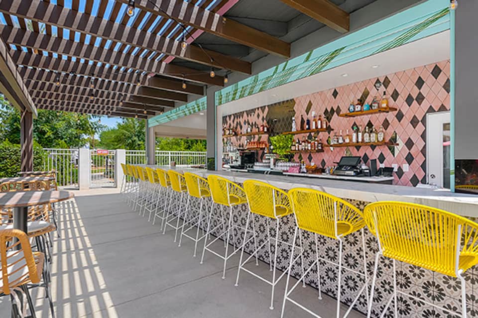 Modern outdoor bar with yellow metal chairs, pergola roof, and pink geometric tile accent wall