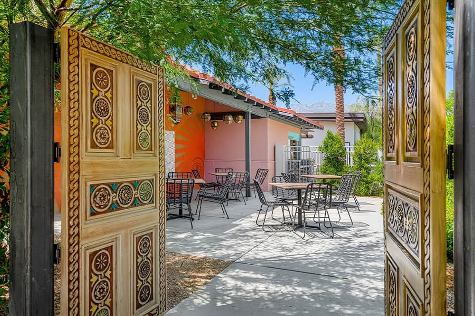 Ornate wooden gates opening to courtyard with coral building, dining tables, and shade structure