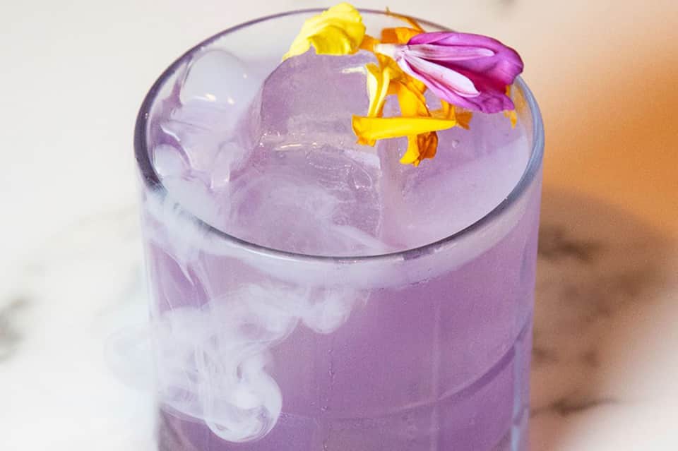Purple cocktail with ice, flowers, and colorful garnish on white surface