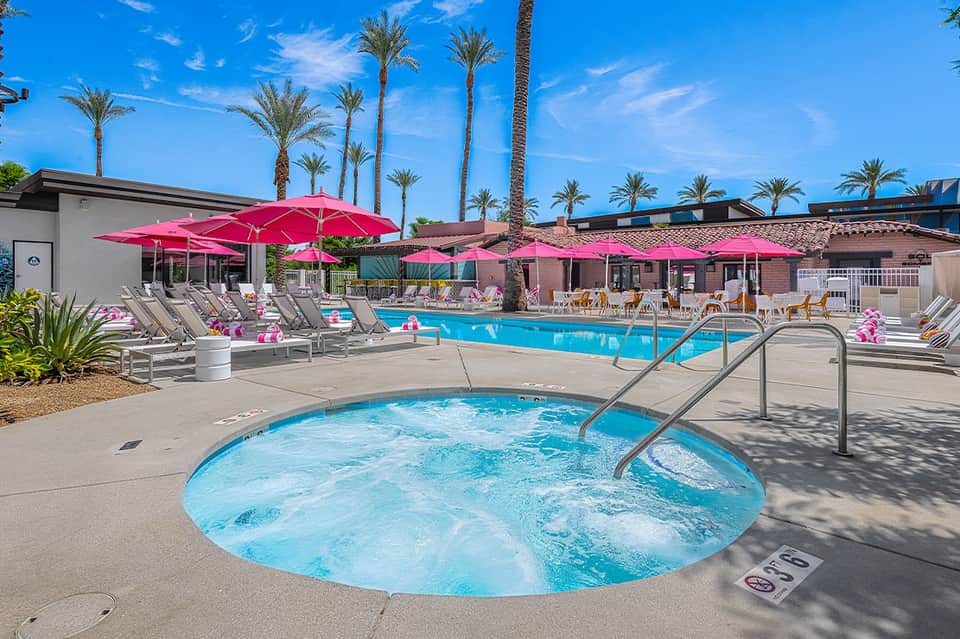 Resort pool complex with hot tub, pink umbrellas, and palm trees under blue sky