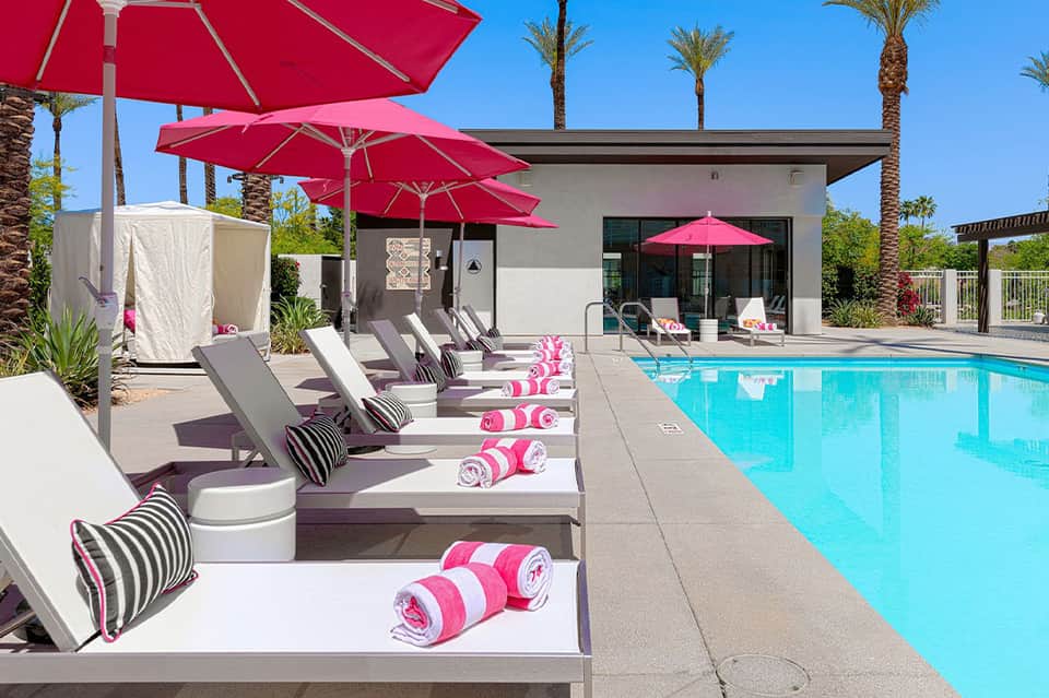 Resort pool area with pink umbrellas, gray lounge chairs, pink towels, and modern spa building under palm trees