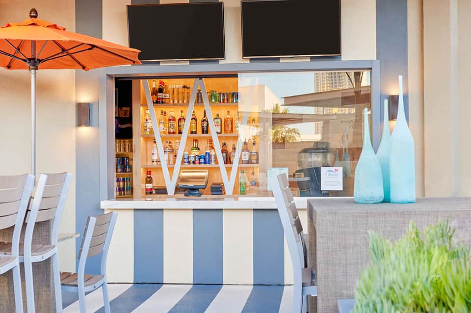 Outdoor bar counter with illuminated liquor display, blue and white striped base, orange umbrella, and seating