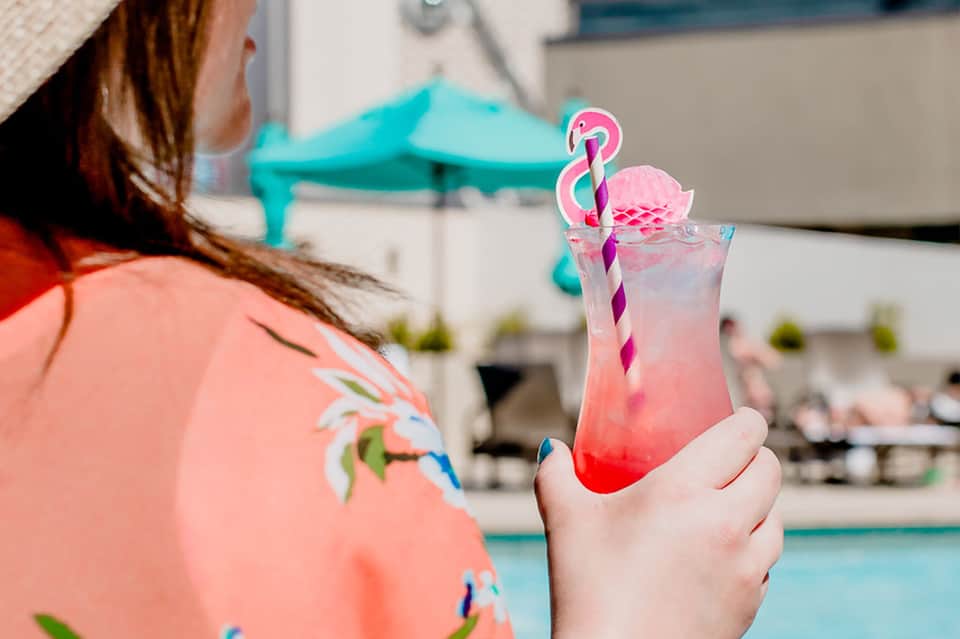 Woman in colorful shirt holding pink layered cocktail with flamingo straw by pool deck