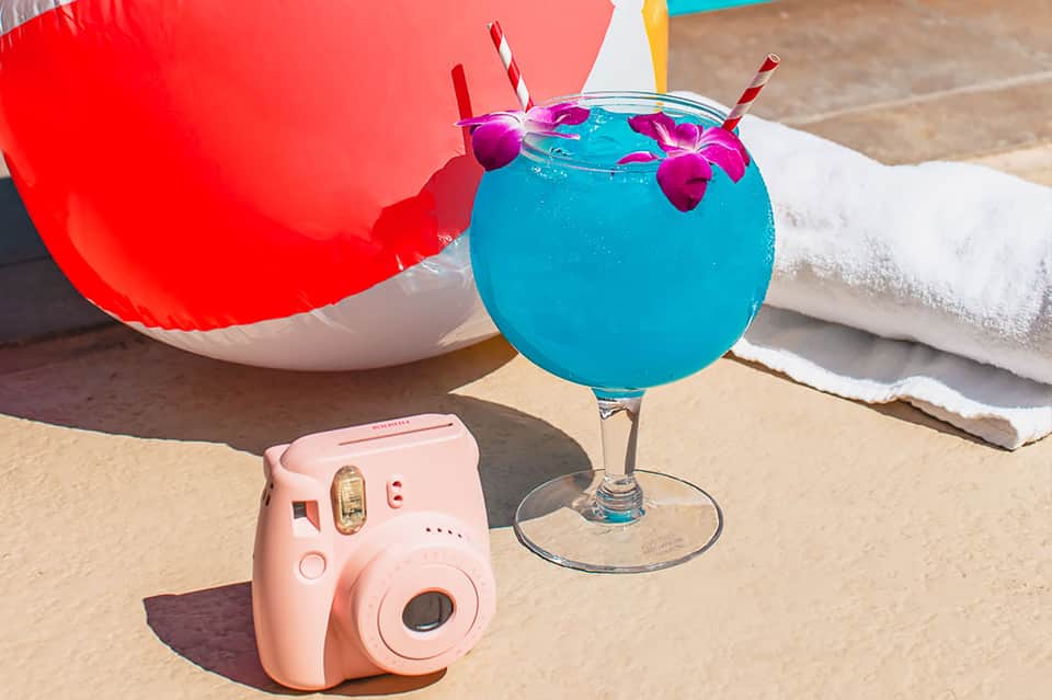 Bright blue tropical cocktail with pink orchid flowers on sandy beach next to pink camera and red beach bag