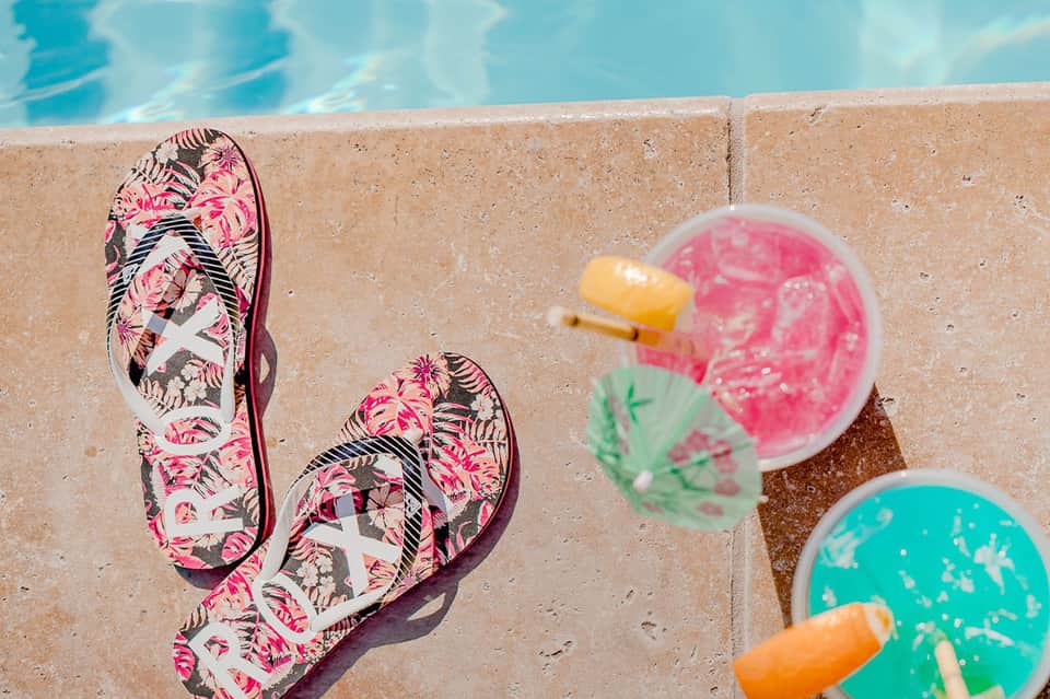 Colorful flip-flops on pool deck next to tropical drinks with umbrellas and pool water