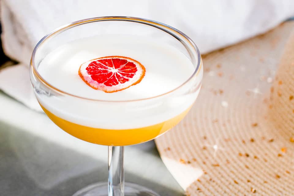 Grapefruit cocktail with foam top and citrus slice garnish on neutral background